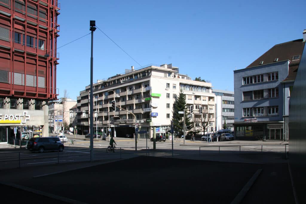 Neubau, Studie, Umbau und Umnutzung von KGRUPPE Architekten Basel - Stadtansicht mit mehreren Gebäuden und einer Strassenkreuzung, links ein rotbraunes Gebäude mit der Aufschrift "Post", im Hintergrund blaues Sommerwetter.