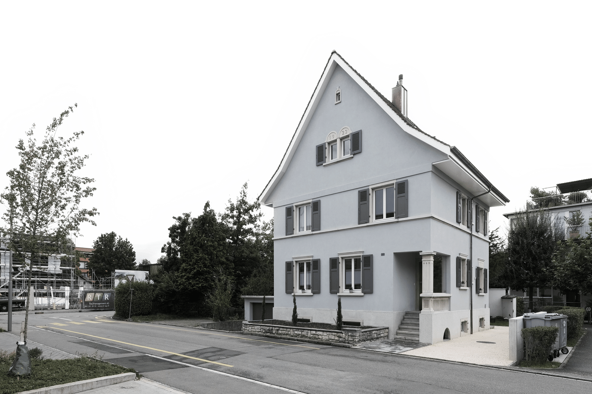 Umbau und Renovation eines Wohnhauses durch das Architekturbüro KGRUPPE Architekten Basel. Ein zweistöckiges Haus mit Satteldach und Fensterläden an den Fenstern, an einer ruhigen Strasse gelegen, mit Bäumen und Gehweg im Vordergrund.