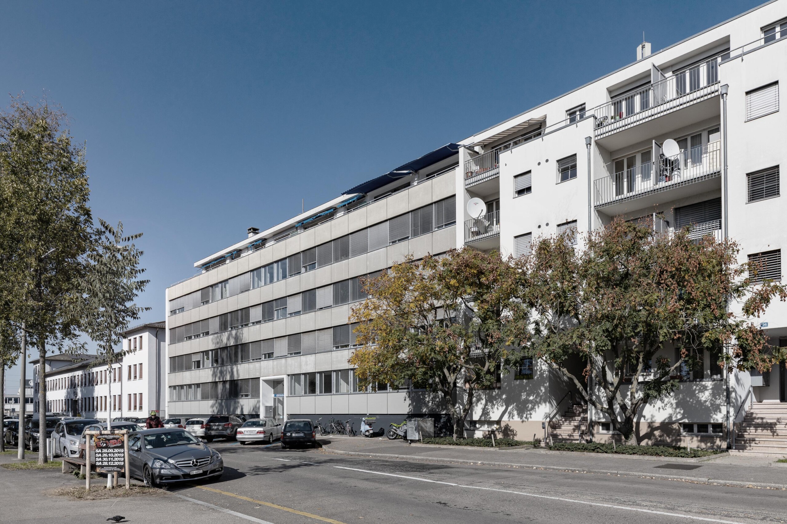 von KGRUPPE Architekten Basel - Bestehendes Bürogebäude an der Hochbergerstrasse 158 mit parkenden Autos und Bäumen im Vordergrund.