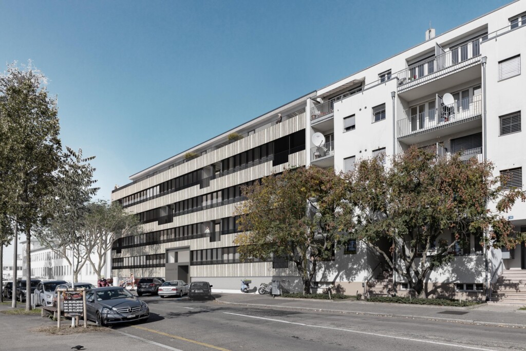 von KGRUPPE Architekten Basel - Umgenutztes, mehrstöckiges Wohngebäude an der Hochbergerstrasse 158 mit modernen Fassaden, Bäumen und parkenden Autos an der Strasse.