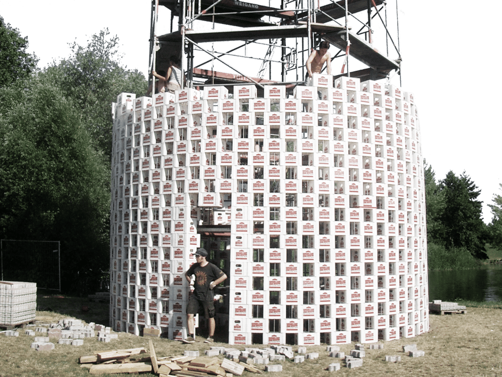 Szenografie, Umnutzung und Upcycling von KGRUPPE Architekten Basel - Ein grosser Turm aus gestapelten Bierkästen im Freien, umgeben von Bäumen und Baugerüsten. Zwei Männer arbeiten auf dem Gerüst, ein weiterer steht am Fusse des Turms.