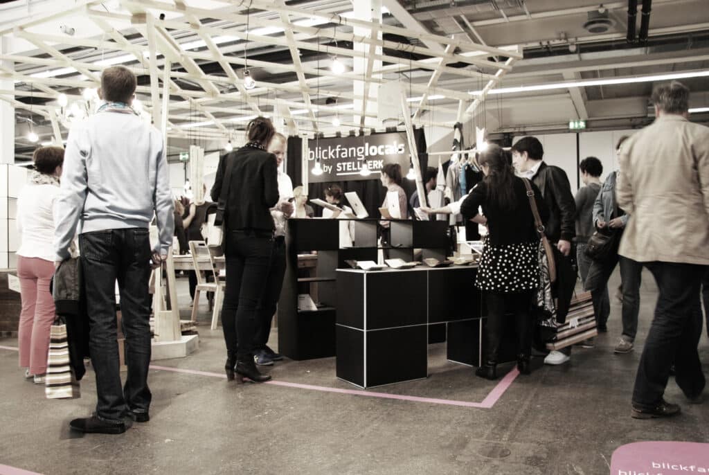 von KGRUPPE Architekten Basel - Menschenmenge an einem Messestand mit der Aufschrift "blickfanglocals by STELLWERK" in einer grossen, beleuchteten Ausstellungshalle.