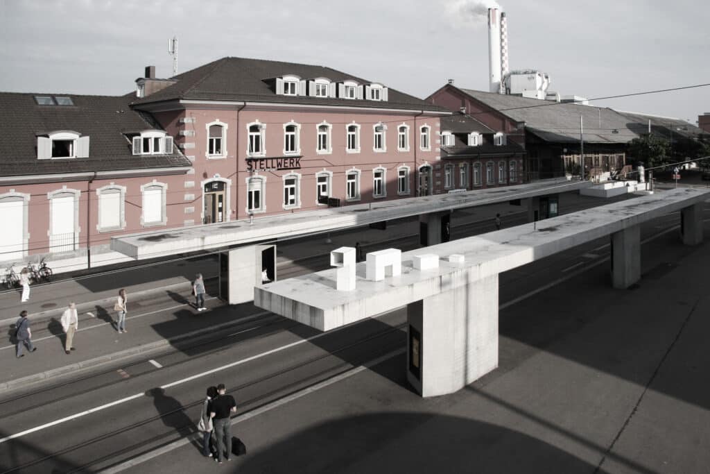 von KGRUPPE Architekten Basel - Bahnhofsgebäude mit dem Schriftzug "Stellwerk", davor moderne Betonplattformen und Gleise, einige Menschen auf dem Bahnsteig.