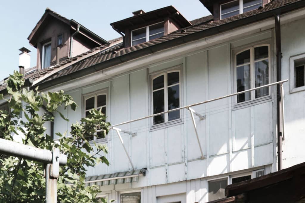 von KGRUPPE Architekten Basel - Aussenansicht eines Wohnhauses mit weissen Holzfassaden, mehreren Fenstern und Dachgauben; im Vordergrund sind dichte grüne Blätter zu sehen.