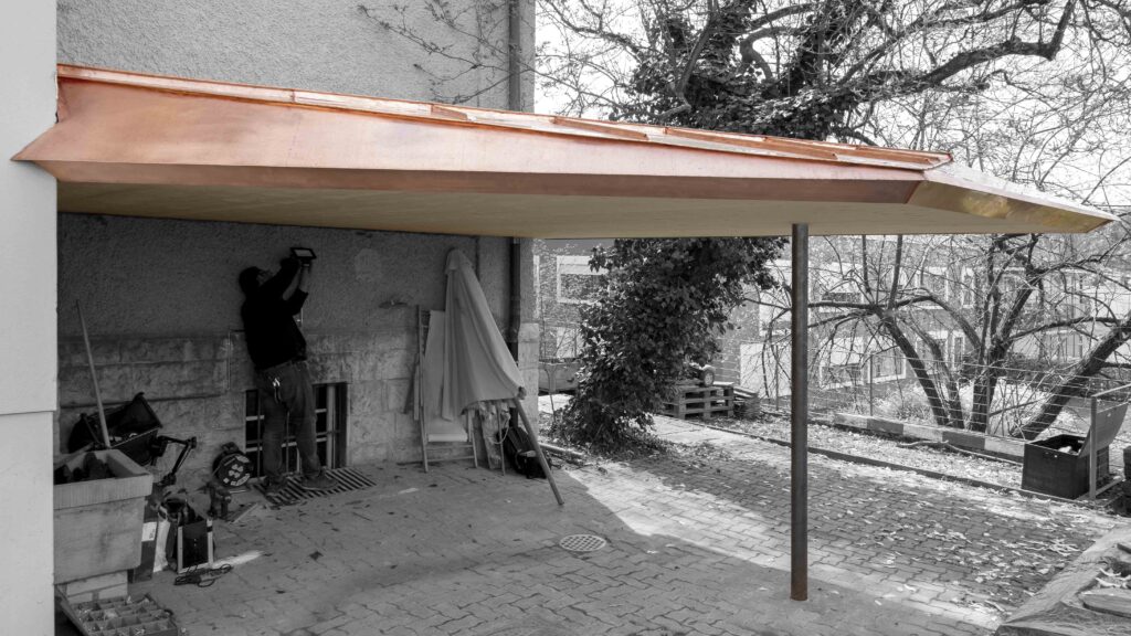 von KGRUPPE Architekten Basel - Ein Handwerker installiert ein Licht unter einem Carport mit Kupferdach, das an einem Haus befestigt ist. Im Hintergrund sind Bäume und eine gepflasterte Auffahrt zu sehen.