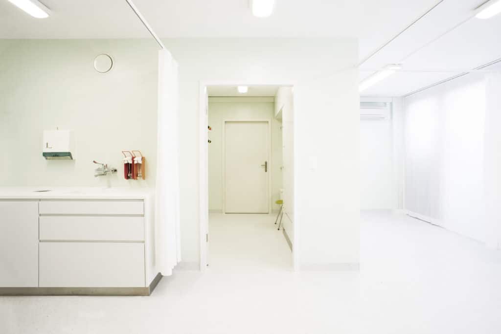 Umbau von KGRUPPE Architekten Basel - Heller Raum in einer Klinik mit weissen Wänden und Möbeln, einem Waschbecken, Seifen- und Desinfektionsmittelspendern sowie einem Durchgang zu einem anderen Zimmer.