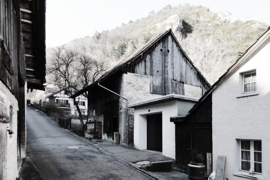 Neubau, Umbau und Umnutzung von KGRUPPE Architekten Basel - Eine enge Strasse in einem Dorf mit traditionellen, alten Häusern und einem bewaldeten Hügel im Hintergrund.