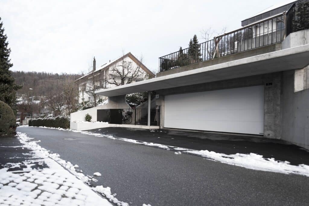 Neubau und Umbau von KGRUPPE Architekten Basel - Moderne Garageneinfahrt mit einem weissen Garagentor, Schnee bedeckt teilweise den Boden und die umliegenden Bäume und Sträucher, im Hintergrund ein Haus und bewaldete Hügel.