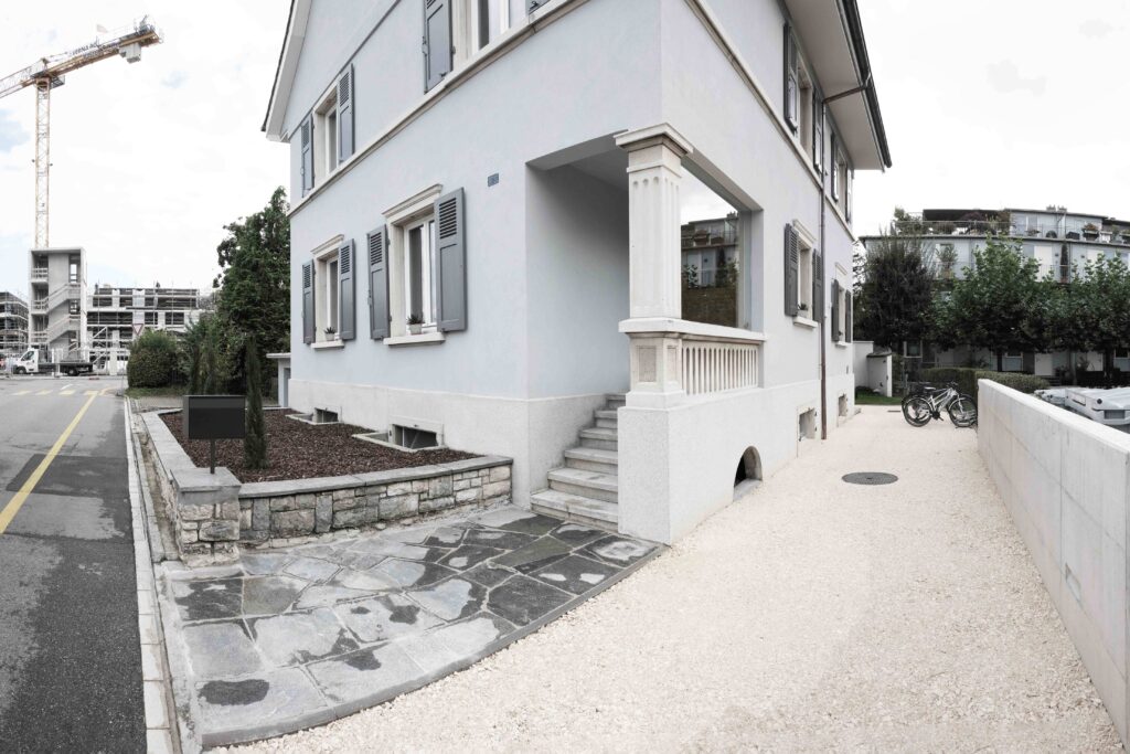 von KGRUPPE Architekten Basel - Eingang eines Gebäudes mit grauer Fassade, Steinmauern und grünen Fensterläden. Baustelle im Hintergrund und Fahrräder an der Seite.