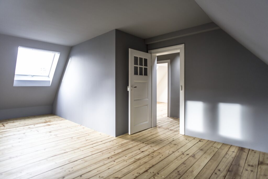 von KGRUPPE Architekten Basel - Modernes Dachgeschosszimmer mit Holzboden, grauen Wänden und einem Dachfenster, Blick durch eine offene Tür in einen angrenzenden Raum.