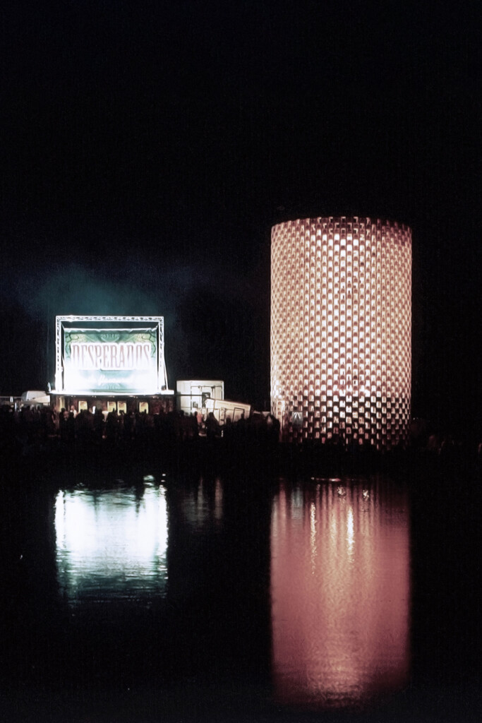 Szenografie, Umnutzung und Upcycling von KGRUPPE Architekten Basel - Nachtaufnahme einer beleuchteten Bühne mit dem Schriftzug "Desperados" und einem hohen zylindrischen Bauwerk, reflektiert im Wasser.