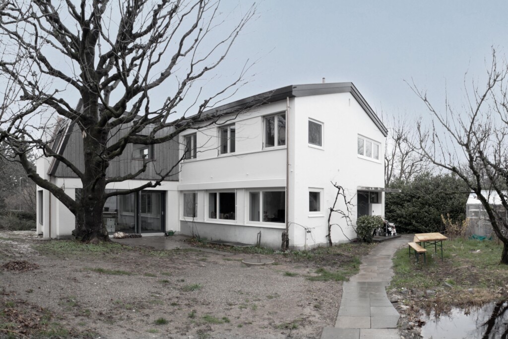 von KGRUPPE Architekten Basel - Umbau und Aufstockung durch KGRUPPE Architekten Basel. Das Foto zeigt das zweistöckige Haus mit grossen Fenstern und steilem Dach, umgeben von kahlen Bäumen und einem Teich im Vordergrund.